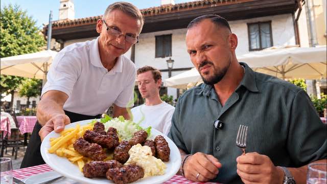 Ein Kellner reicht einen Teller mit Ćevapi und Pommes, am Tisch verzieht ein Gast das Gesicht.